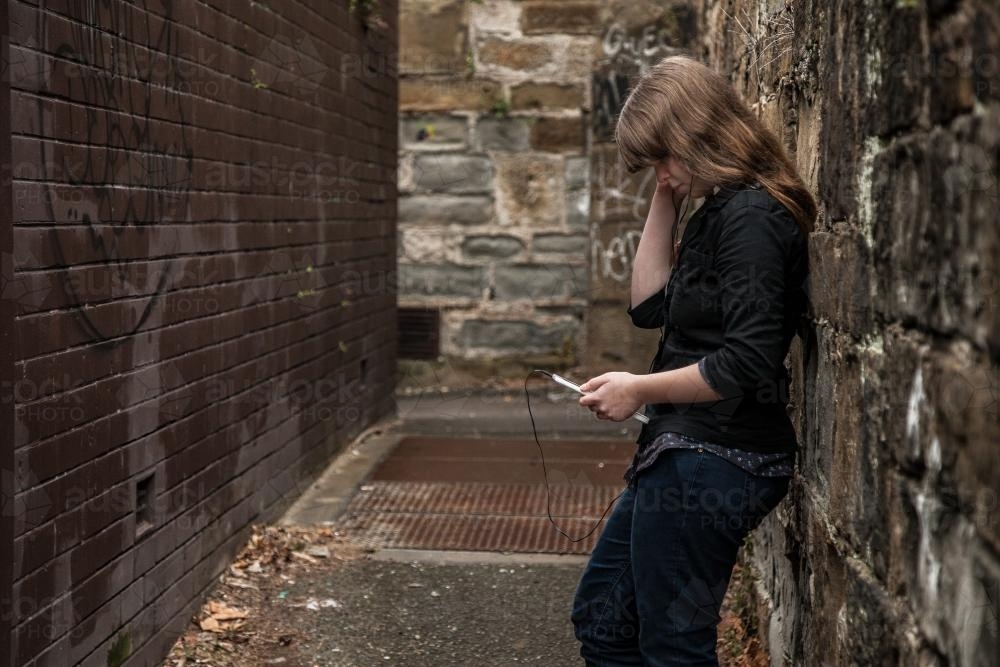 Young teen girl leaning on stone alley wall listening to music - Australian Stock Image