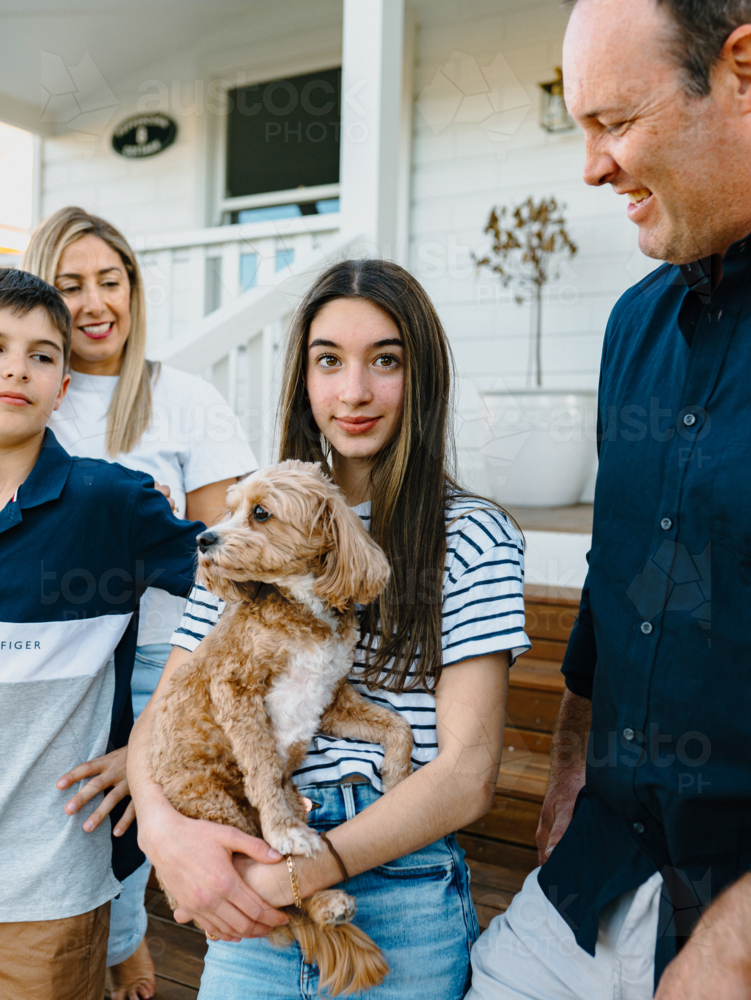 Young teenage girl carrying a dog while standing beside her parents and brother outside the house. - Australian Stock Image