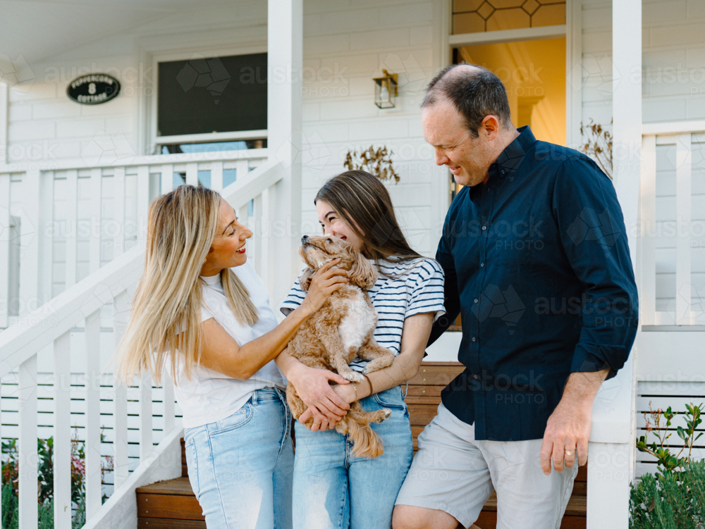 Young teenage girl carrying a dog while standing beside her parents outside the house. - Australian Stock Image