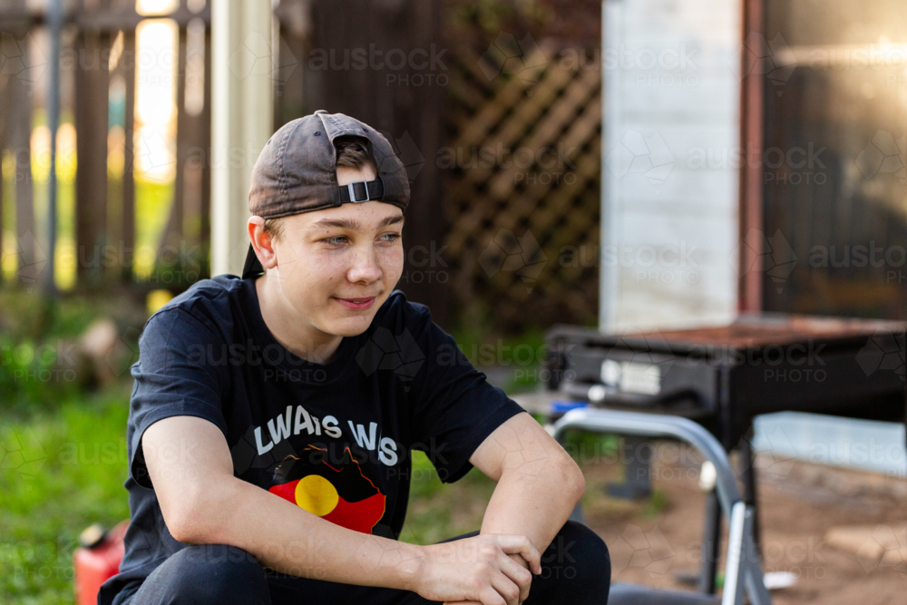 Young teenaged Aboriginal boy sitting in backyard of home wearing cap backwards - Australian Stock Image