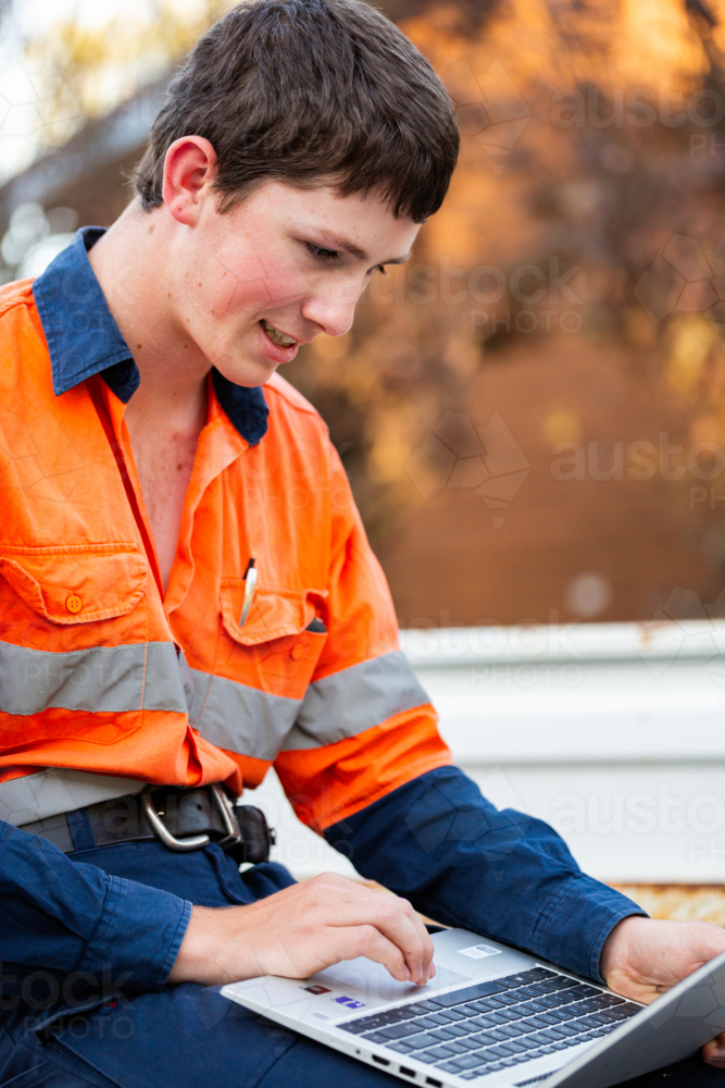 Young teenaged trainee tradie apprentice working on laptop in back of ute in high-vis workwear - Australian Stock Image