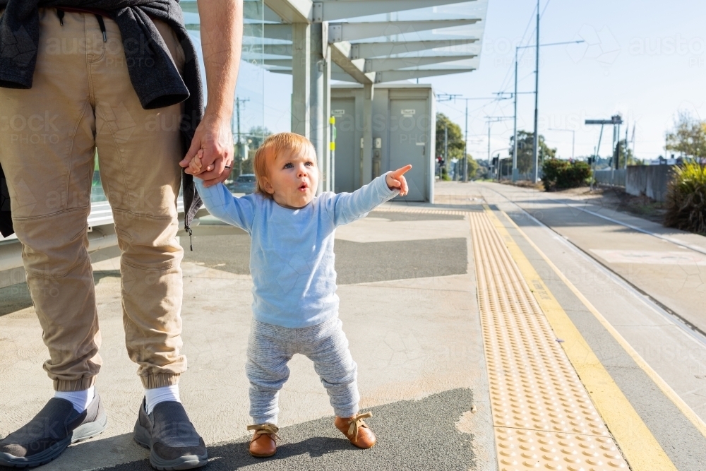 Young toddler pointing at tram station platform holding parents hand - Australian Stock Image