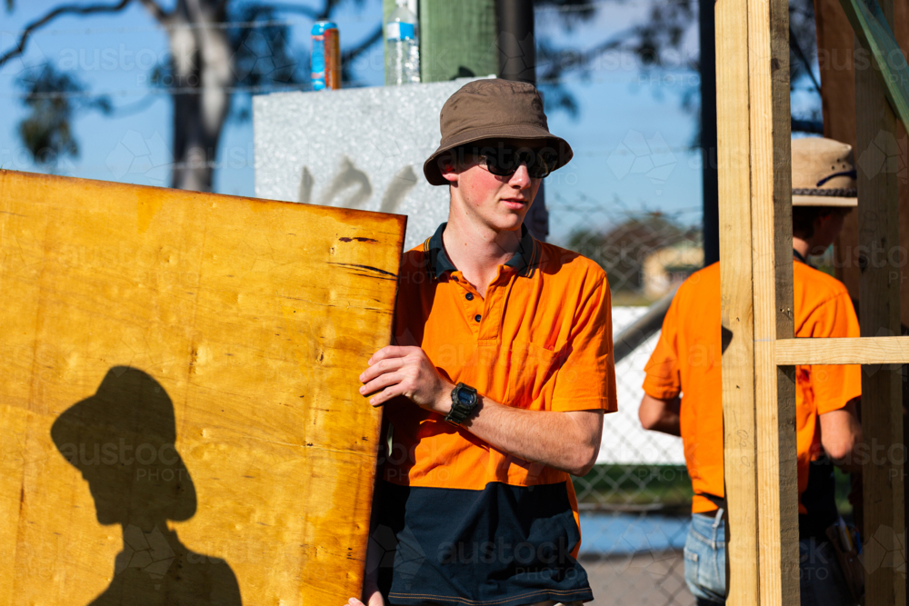 young tradesman carrying sheet of wood towards building frame - Australian Stock Image