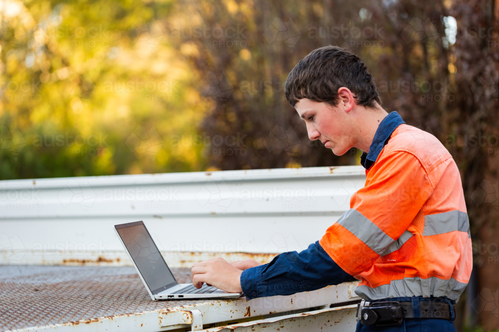 Young trainee tradie apprentice working on laptop in back of ute in high-vis workwear - Australian Stock Image