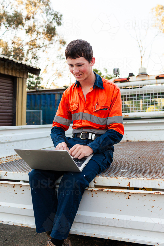 Young trainee tradie apprentice working on laptop in back of ute in high-vis workwear - Australian Stock Image