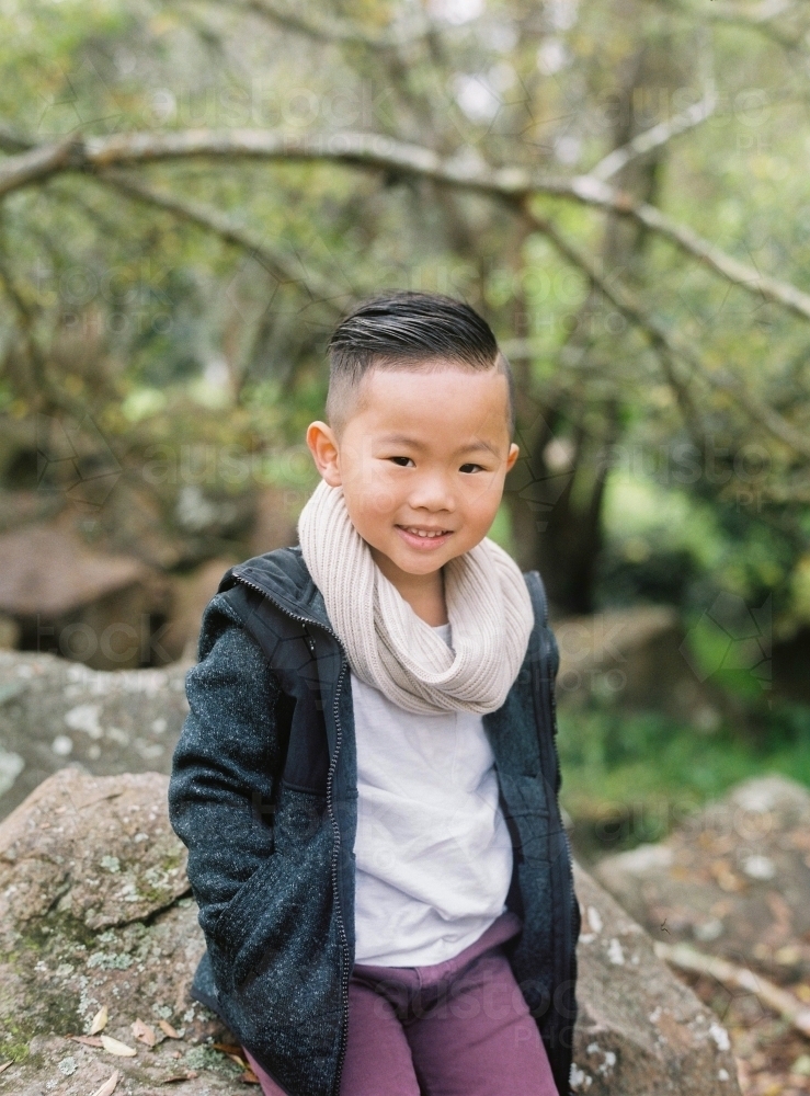 Young, trendy, Asian boy sitting on rock, smiling at the camera - Australian Stock Image