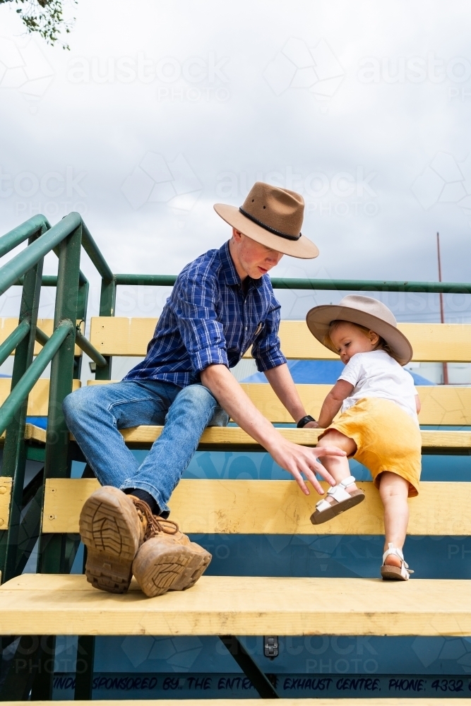 Young uncle sitting on grandstand seats with his toddler niece at local agricultural show - Australian Stock Image