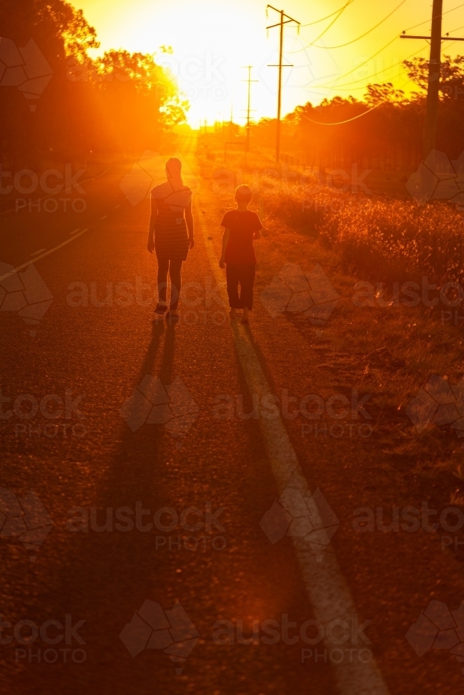 Young woman and young boy walking along the side of the road towards the sunset - Australian Stock Image