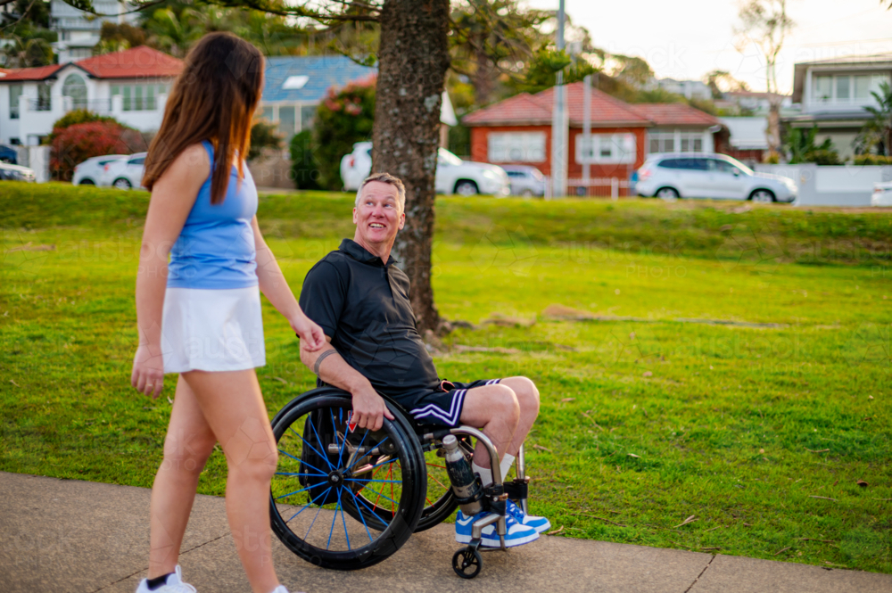 Young woman approaches smiling man in wheelchair, sharing a moment in afternoon sunlight at park - Australian Stock Image