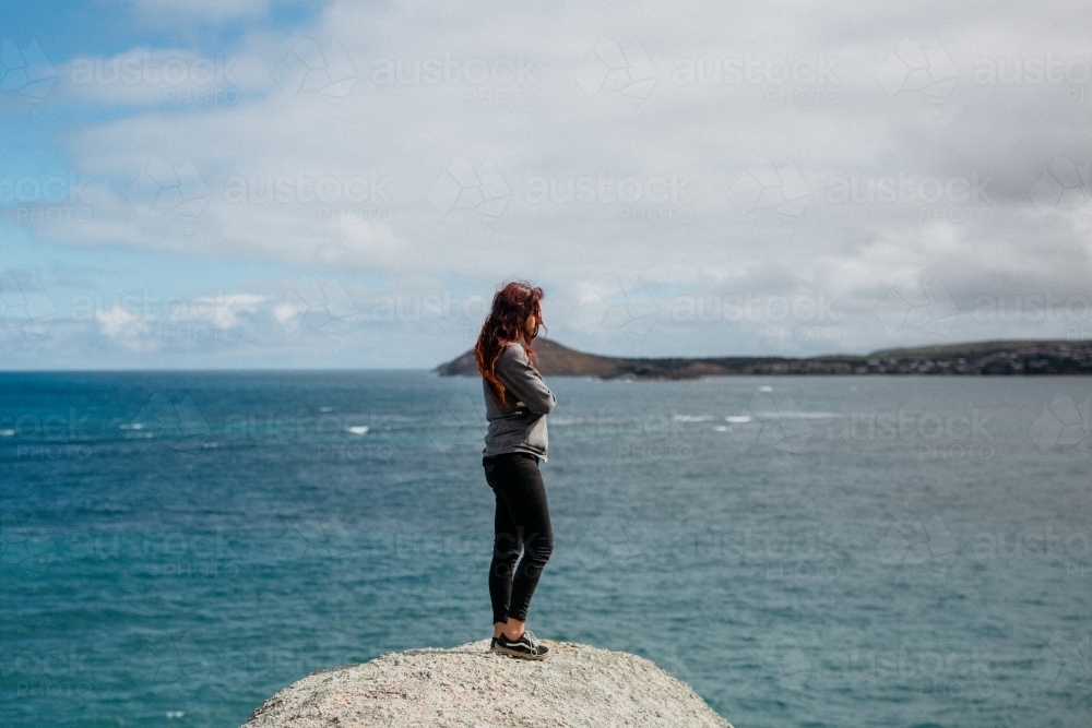 Young woman arms crossed standing on rock looking at ocean - Australian Stock Image