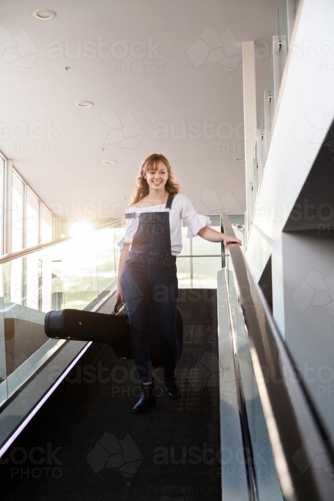 Young woman carrying guitar case down escalator in shopping center - Australian Stock Image