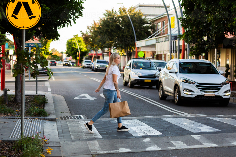 young woman crossing street on pedestrian crossing in urban area of country town carrying shopping - Australian Stock Image