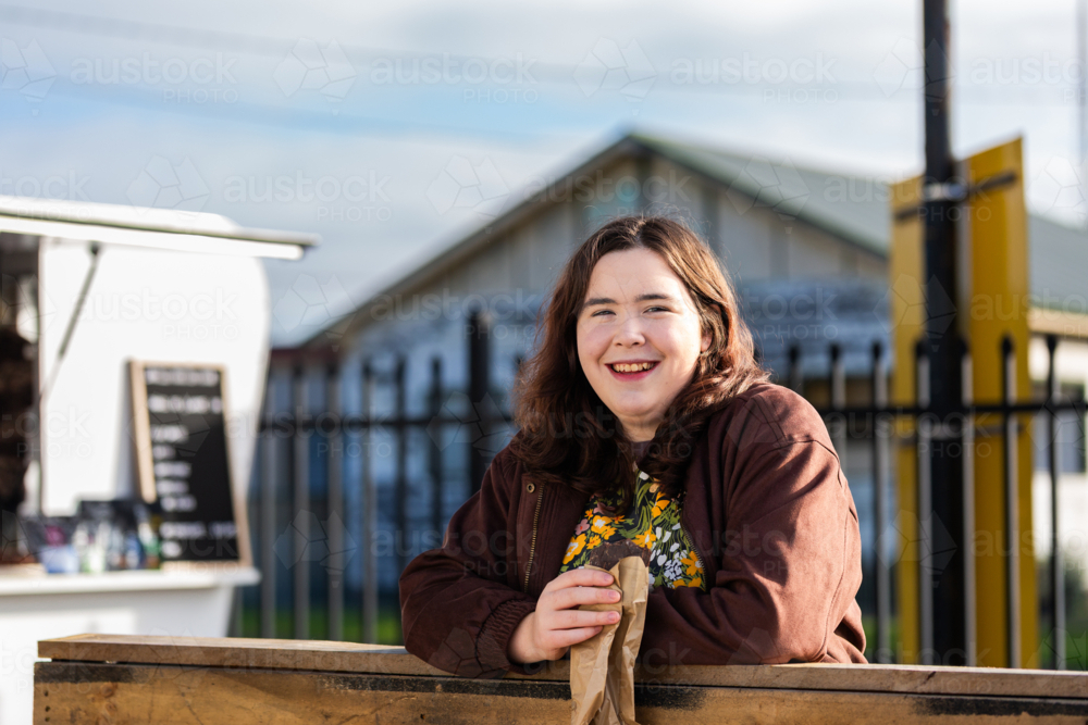Young woman eating brownie treat in brown paper bag from food truck in morning light - Australian Stock Image
