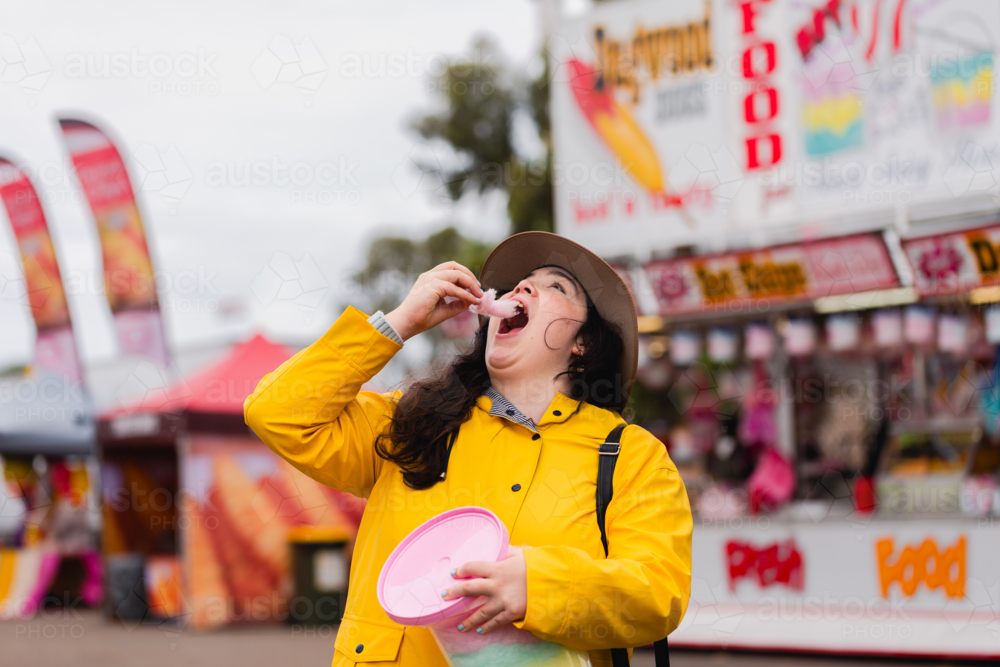 Young woman eating fairy floss from bucket at country show on overcast day in yellow rain jacket - Australian Stock Image