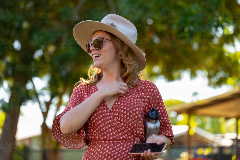 Young woman going to event wearing hat and sunglasses, carrying phone and water bottle - Australian Stock Image