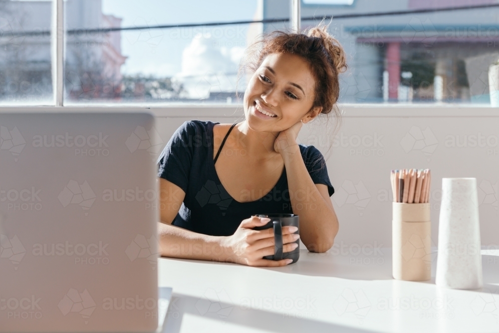 Young woman having coffee at a desk smiling at her computer screen - Australian Stock Image