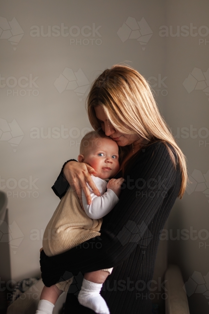 Young woman hugging her baby boy side lit by window of home - Australian Stock Image