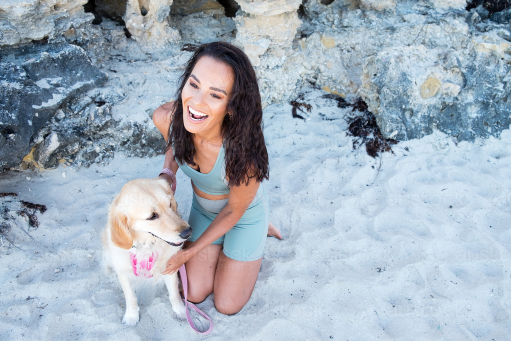 Young woman in active wear kneeling next to large golden dog on beach - Australian Stock Image