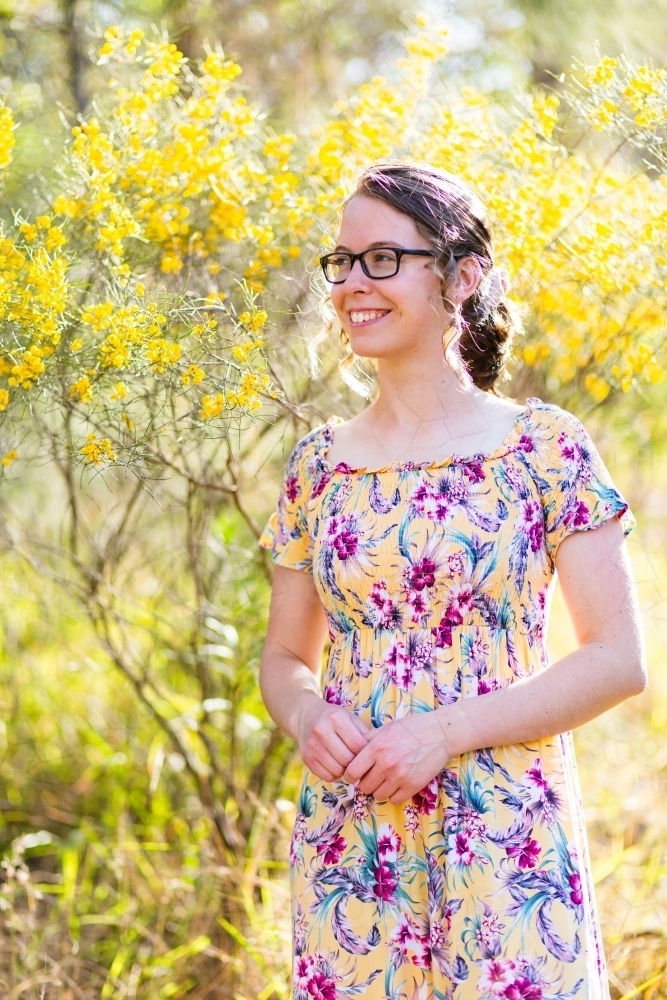 Young woman in bright floral dress standing near yellow flowering bush - Australian Stock Image