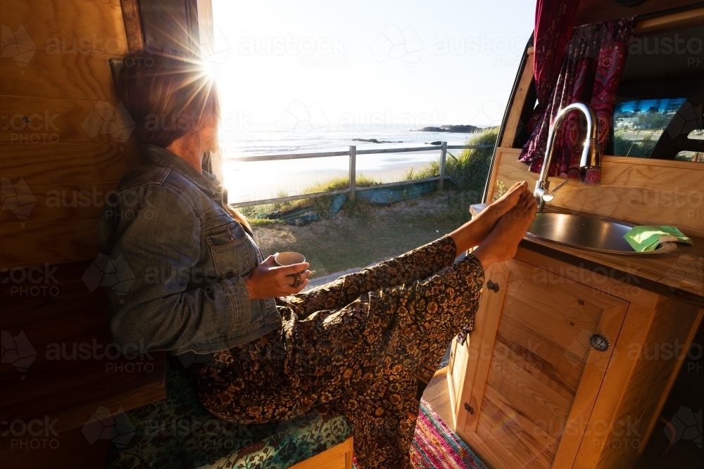 Young woman in camper van watching summer sunrise while drinking coffee by the beach - Australian Stock Image