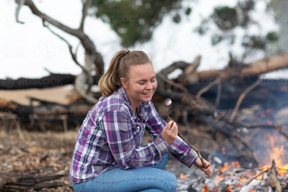 Young woman in checked shirt and jeans toasting marshmallows on campfire - Australian Stock Image