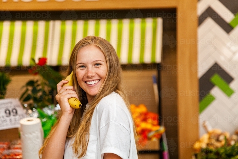 Young woman in grocery store holding banana to her ear - Australian Stock Image
