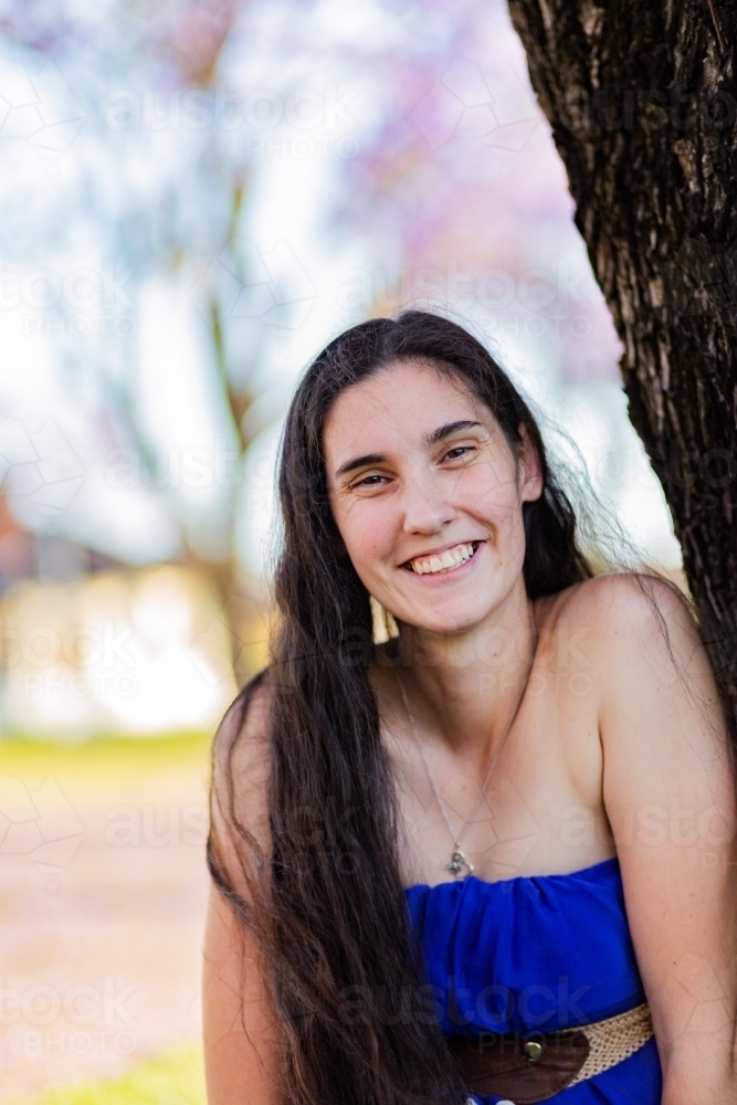 Young woman in her twenties smiling outside leaning on tree - Australian Stock Image