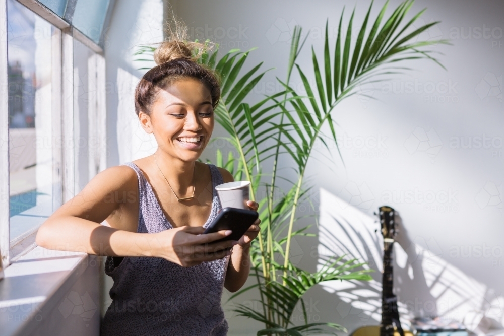 Young woman laughing while having coffee and looking at her phone - Australian Stock Image