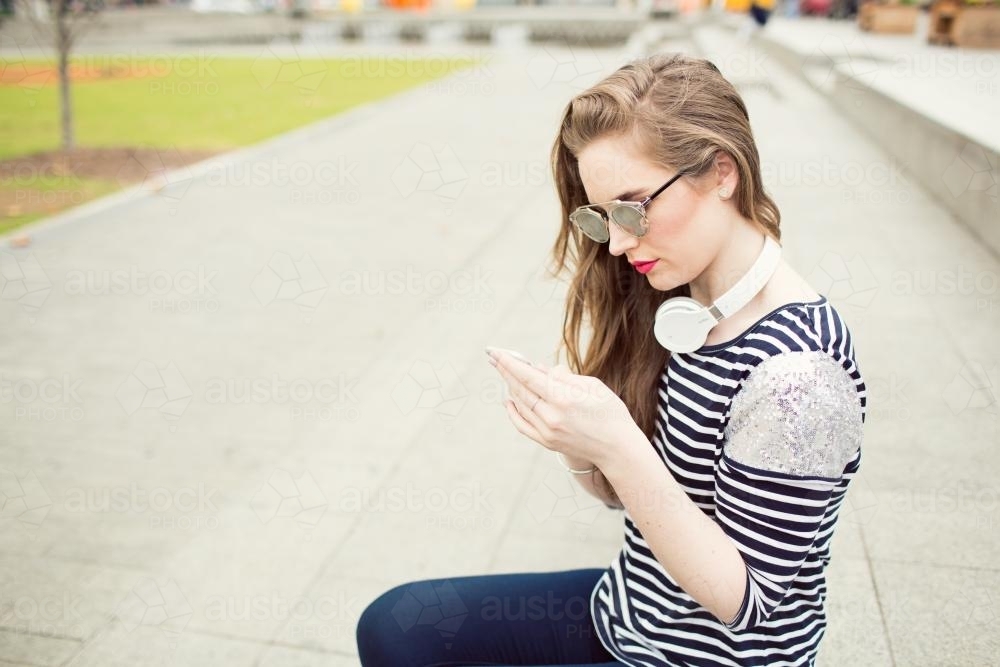 Young woman looking at phone wearing sunglasses and headphones - Australian Stock Image