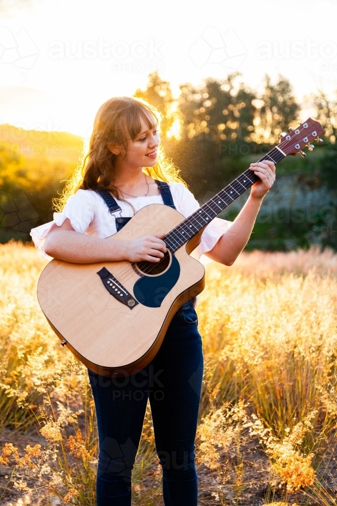 Young woman playing guitar in grass backlit by golden light - Australian Stock Image