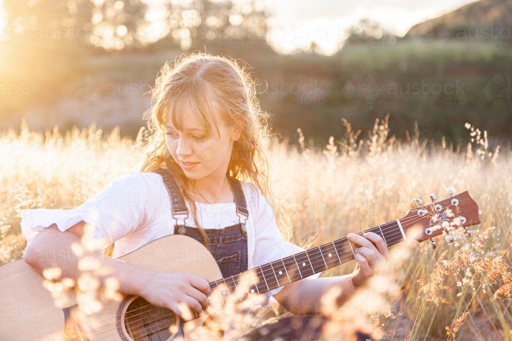 Young woman playing guitar in grass backlit by golden light - Australian Stock Image