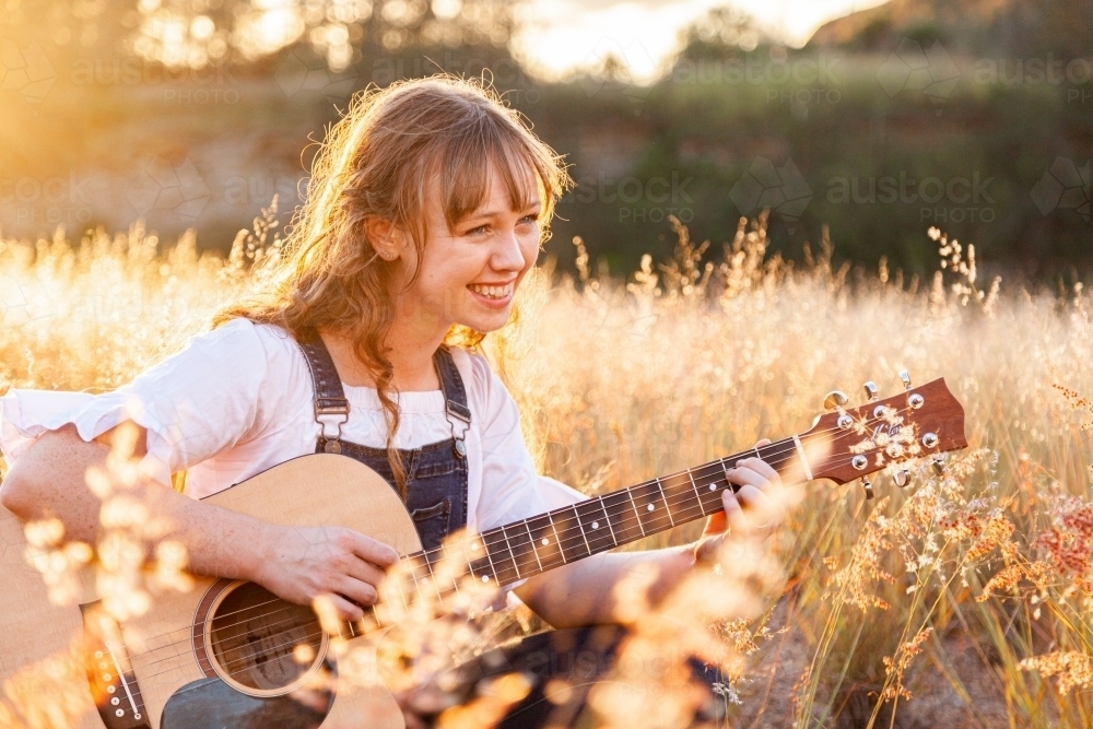 Young woman playing guitar in grass backlit by golden light - Australian Stock Image