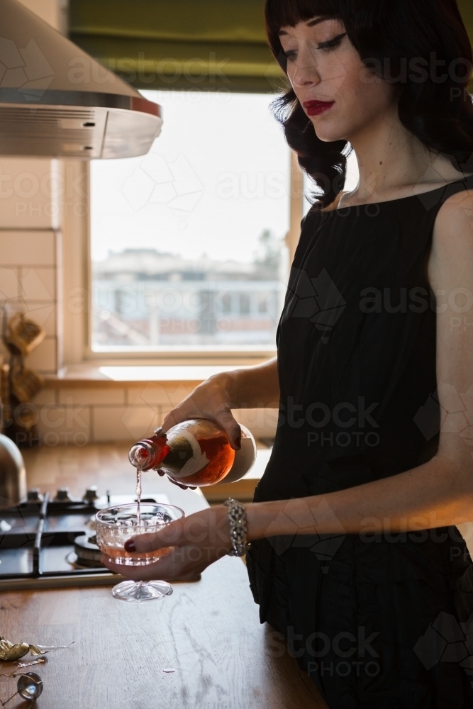 Young woman pouring a glass of pink champagne in the kitchen - Australian Stock Image