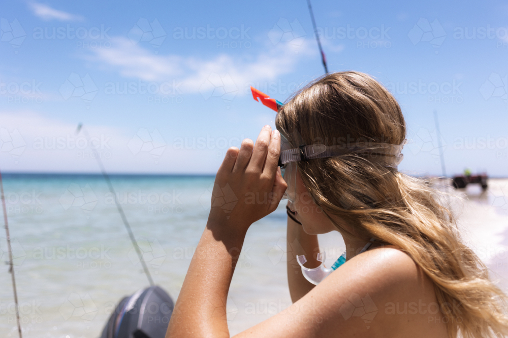 Young woman putting on snorkelling equipment at beach with boat in background - Australian Stock Image