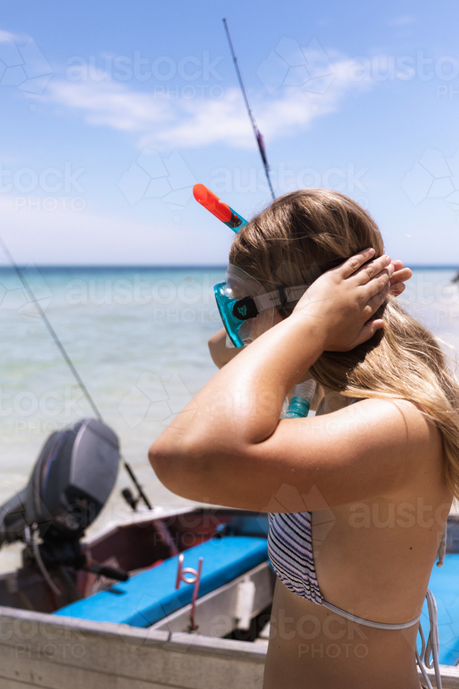 Young woman putting on snorkelling gear at the beach with small boat in background - Australian Stock Image