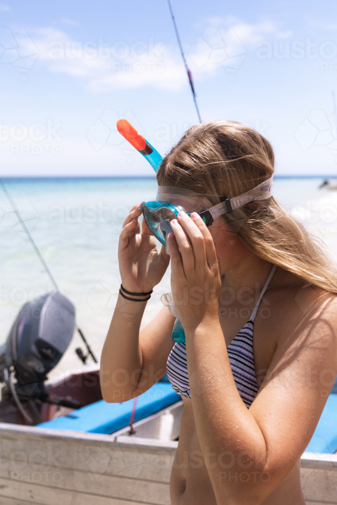 Young woman putting on snorkelling mask at beach with boat in background - Australian Stock Image