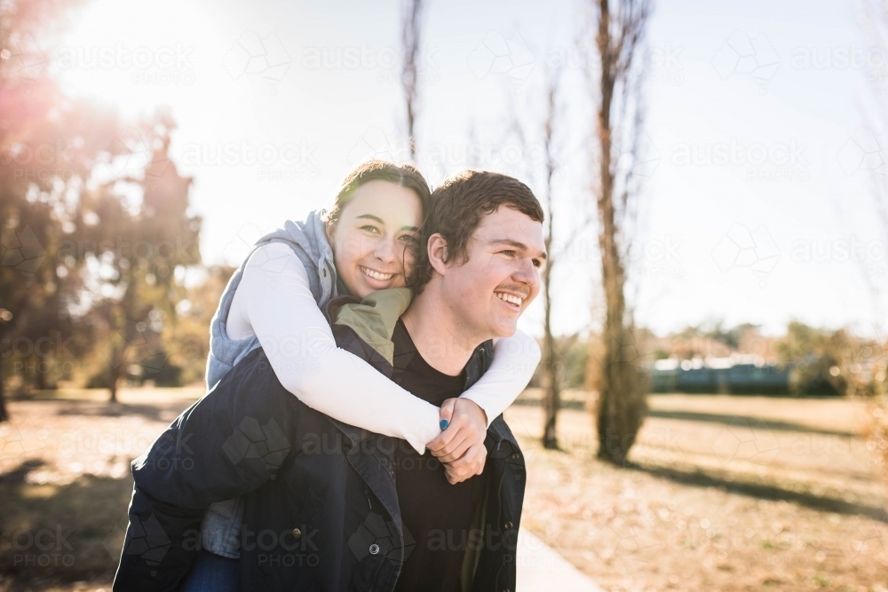Young woman riding piggy back on boyfriend with arms around neck smiling - Australian Stock Image