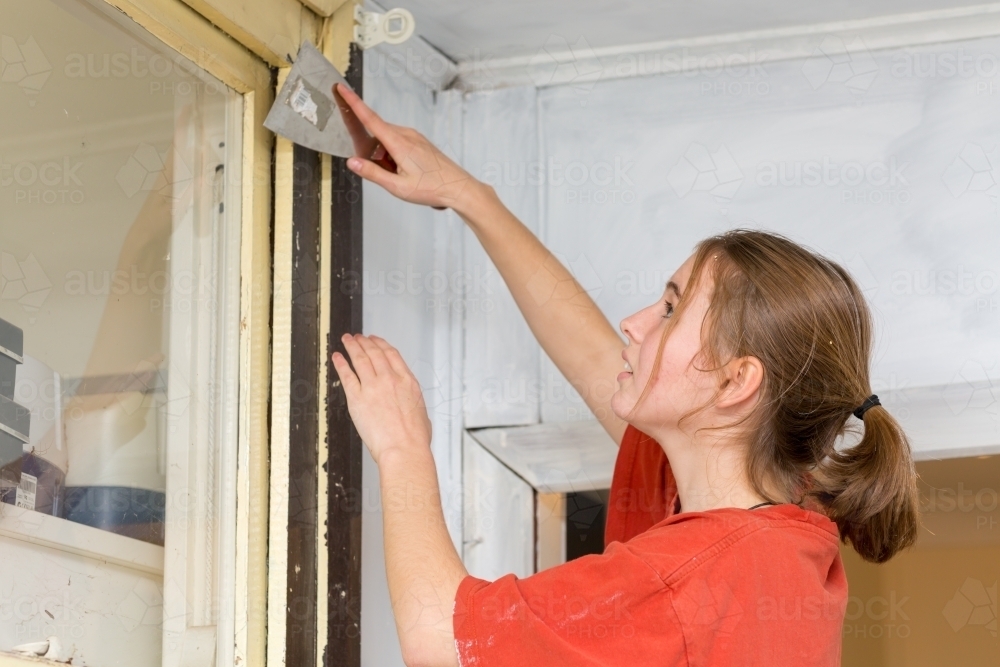 Young woman scraping paint off window frame - Australian Stock Image