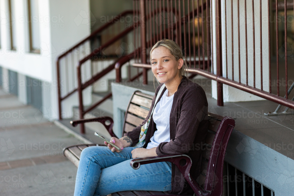young woman seated on park bench chair in urban area of country town with phone - Australian Stock Image