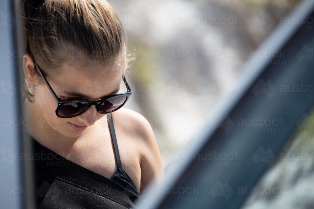 Young woman seen through open door of car with sunglasses and hair tied back - Australian Stock Image