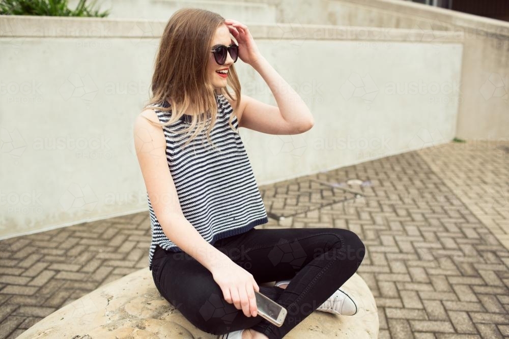 Young woman sitting against a wall with a phone and sunglasses - Australian Stock Image