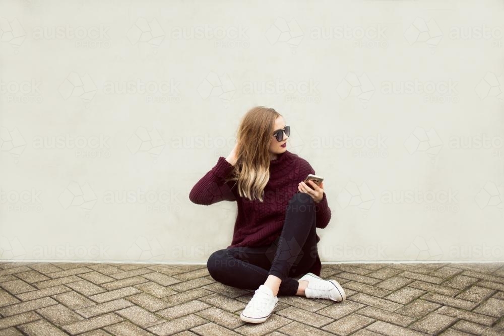 Young woman sitting against a wall with a phone and sunglasses - Australian Stock Image