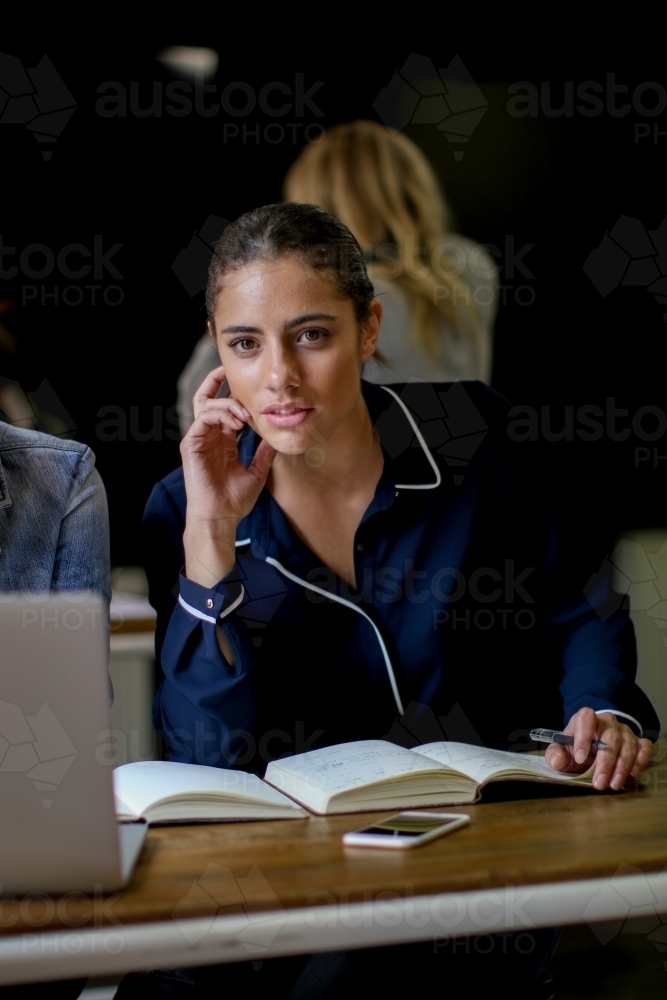 Young woman sitting at desk with book and laptop staring straight at the camera - Australian Stock Image