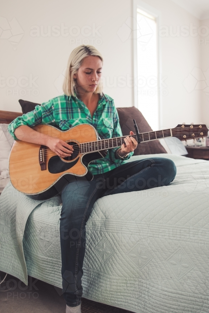 Young woman sitting on a bed looking down as she plays the guitar - Australian Stock Image