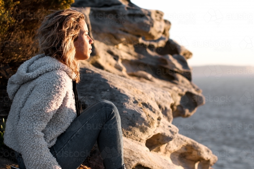 Young woman sitting on a clifftop looking out at the ocean at sunrise : Austockphoto Young woman sitting on a clifftop looking out at the ocean at sunrise - Australian Stock Image
