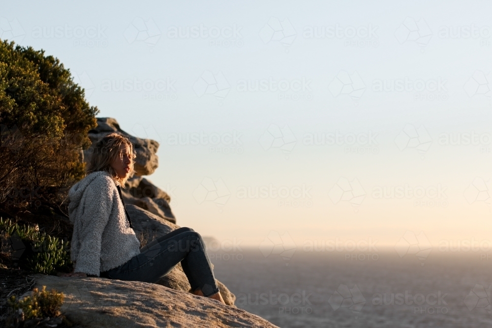 Young woman sitting on a clifftop looking out at the ocean at sunrise - Australian Stock Image