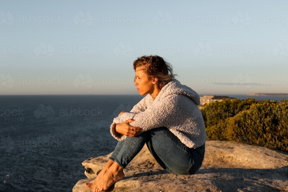 Young woman sitting on a clifftop looking out at the ocean at sunrise - Australian Stock Image