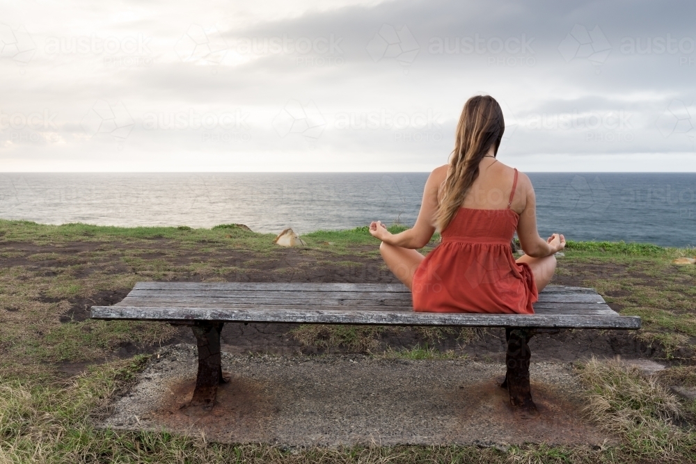 Young woman sitting on a park bench looking out to sea in a meditation pose - Australian Stock Image
