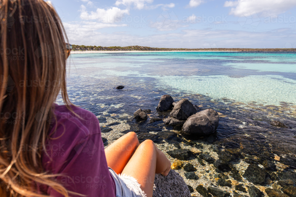 Young woman sitting on a rock beside the sea with view of pristine beach and coastline in summer - Australian Stock Image