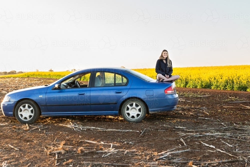 Young woman sitting on boot of car in dirt paddock near canola field on farm - Australian Stock Image